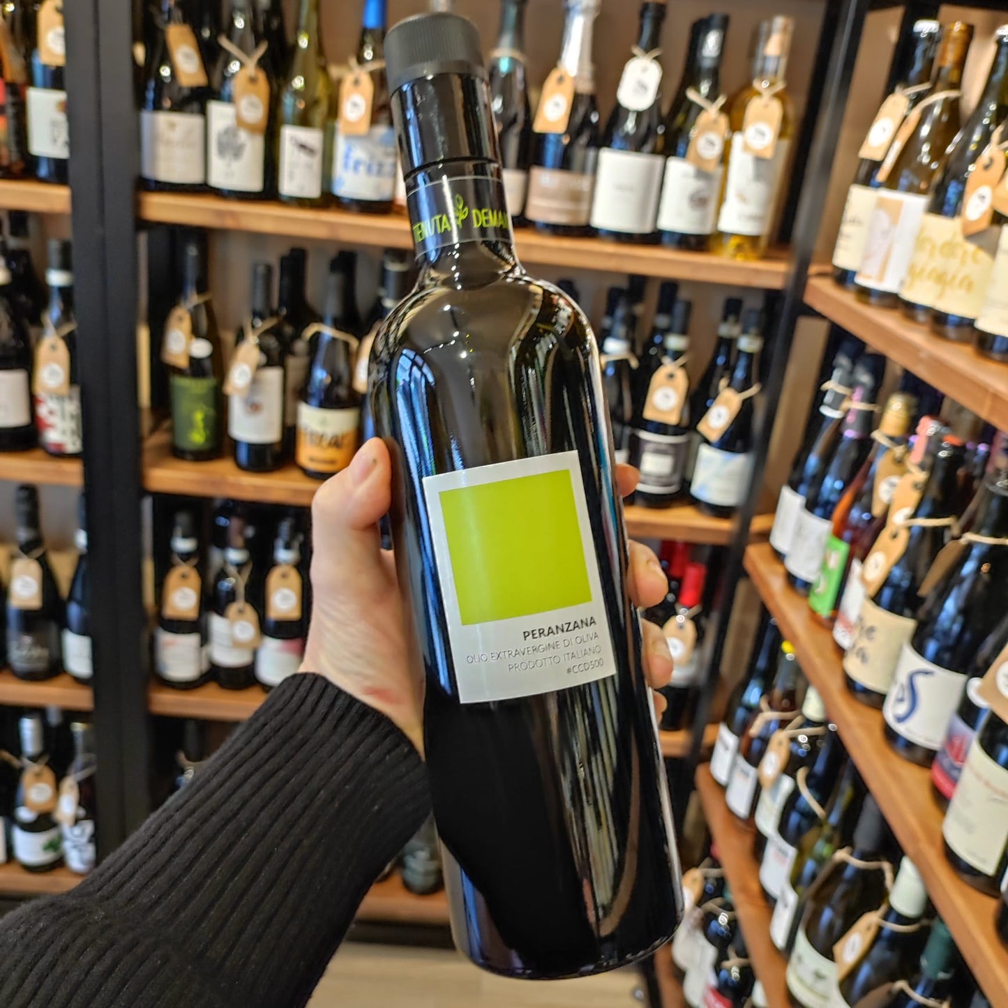 Bottiglia di olio extravergine di oliva Peranzana tenuta in mano in un negozio specializzato, circondata da bottiglie di vini italiani.
