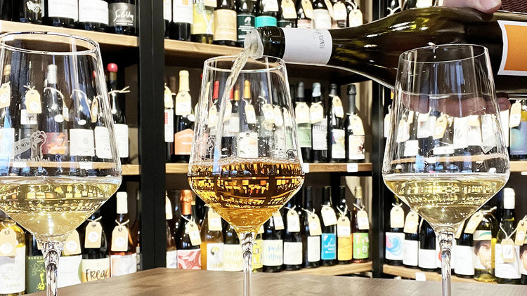 Degustazione di vini bianchi naturali e orange wine in enoteca, con calici riempiti a mano e scaffali di bottiglie artigianali sullo sfondo